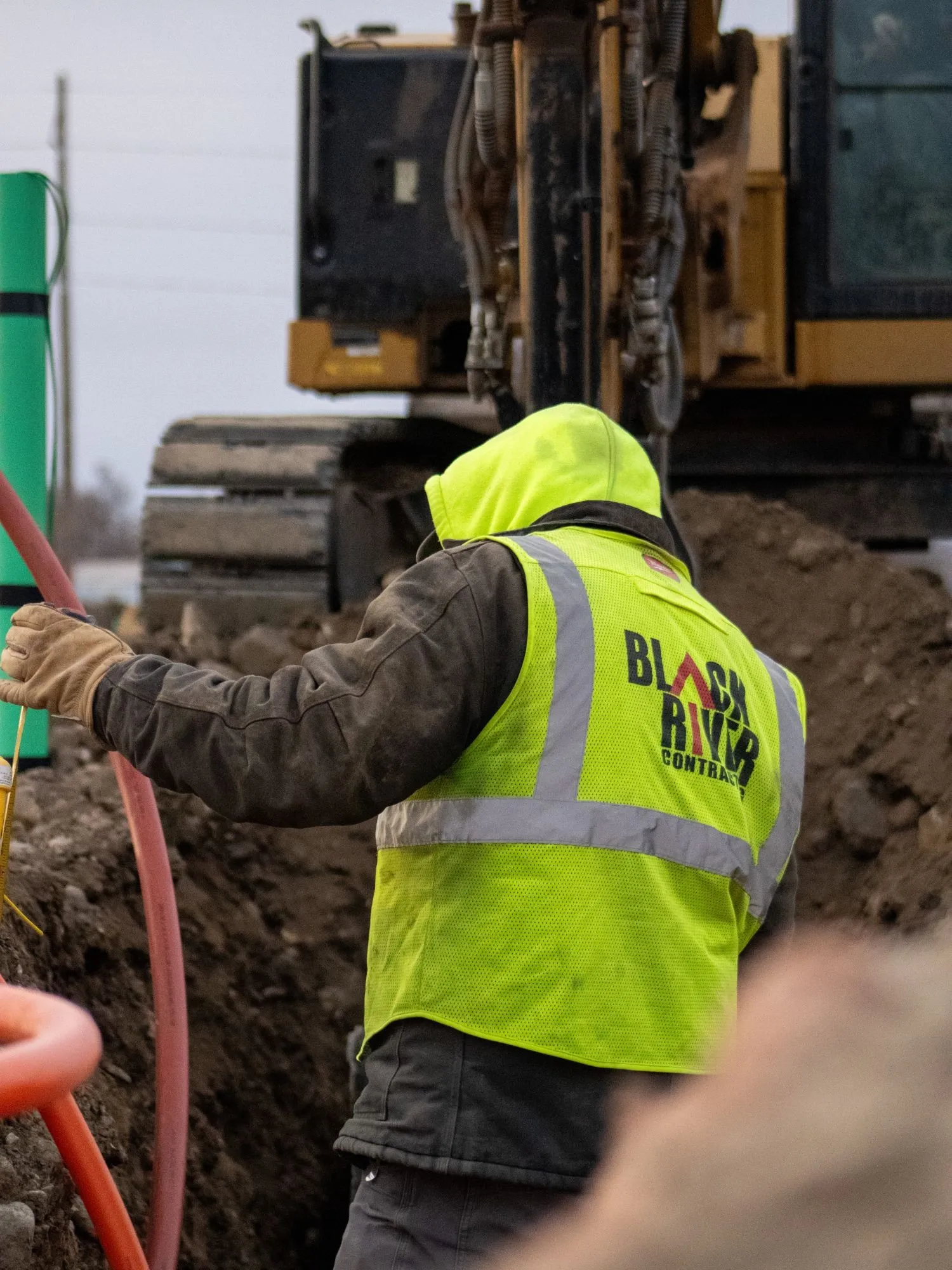 A Black River crew member guiding conduit into a trench with a Cat 309 excavator working behind