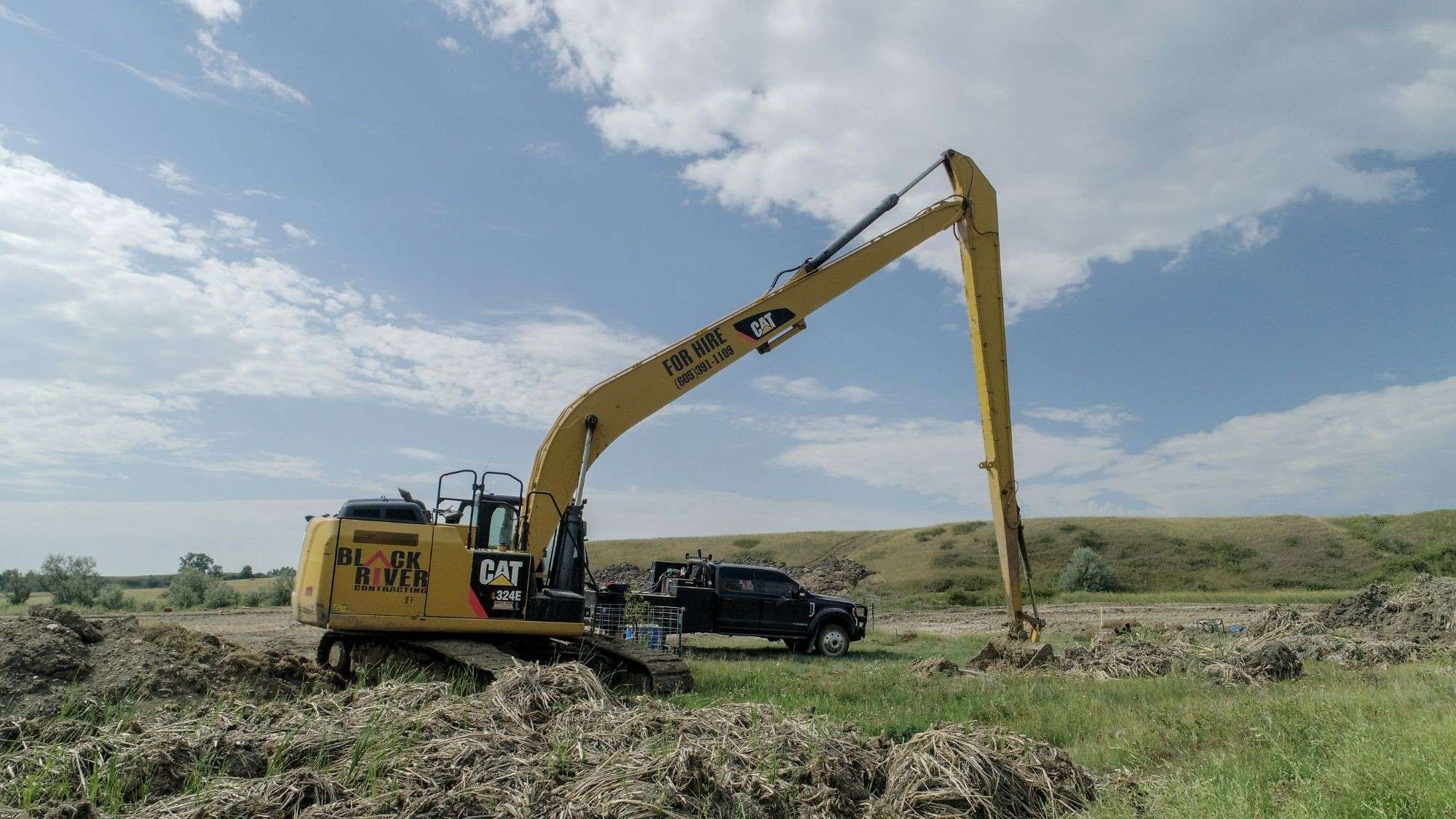 Black River Cat 324E long-reach excavator with FOR HIRE on the boom, beside a pond cleanout