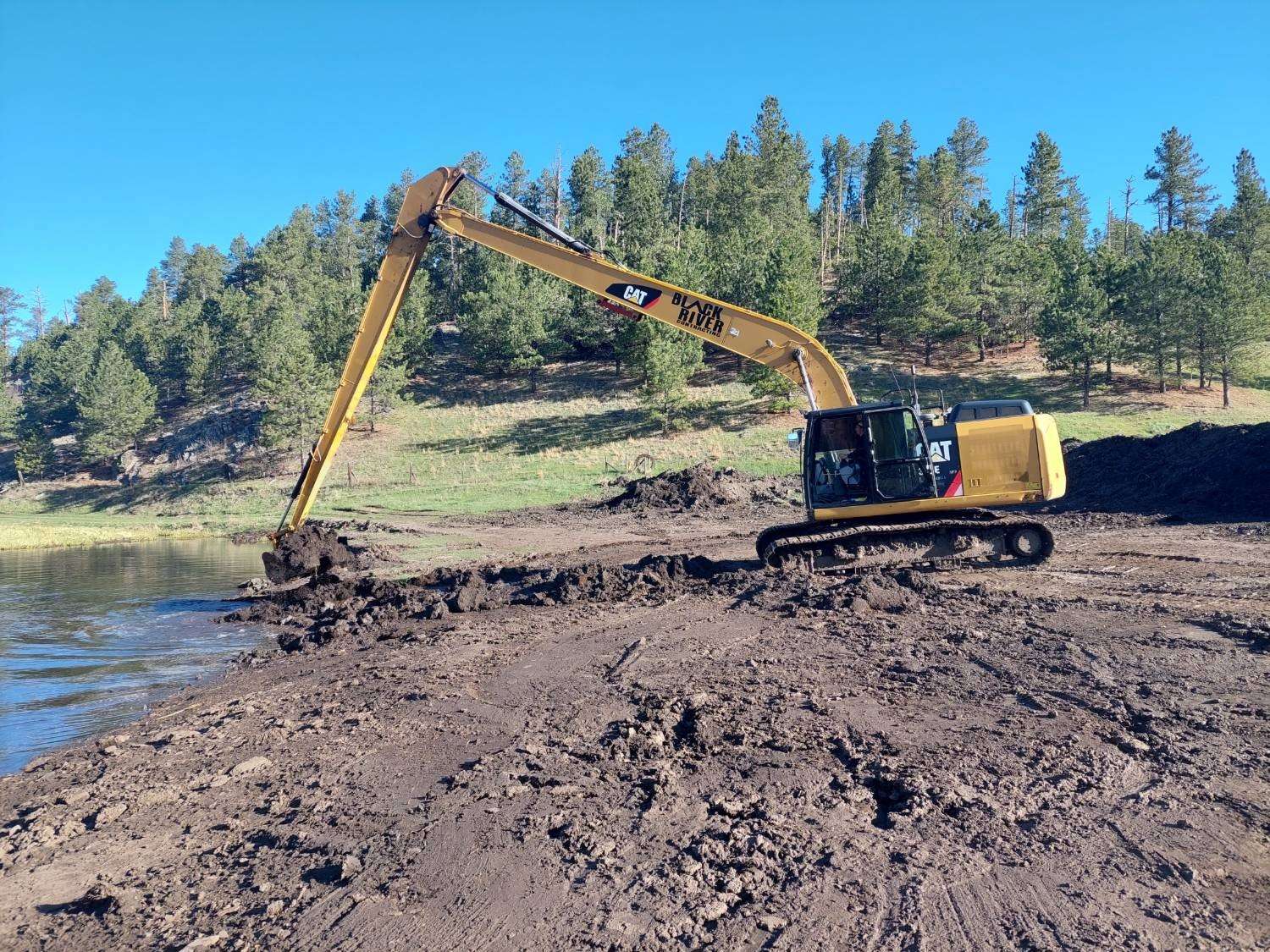 Black River Cat long-reach excavator pulling silt from the edge of a pond below pine-covered hills