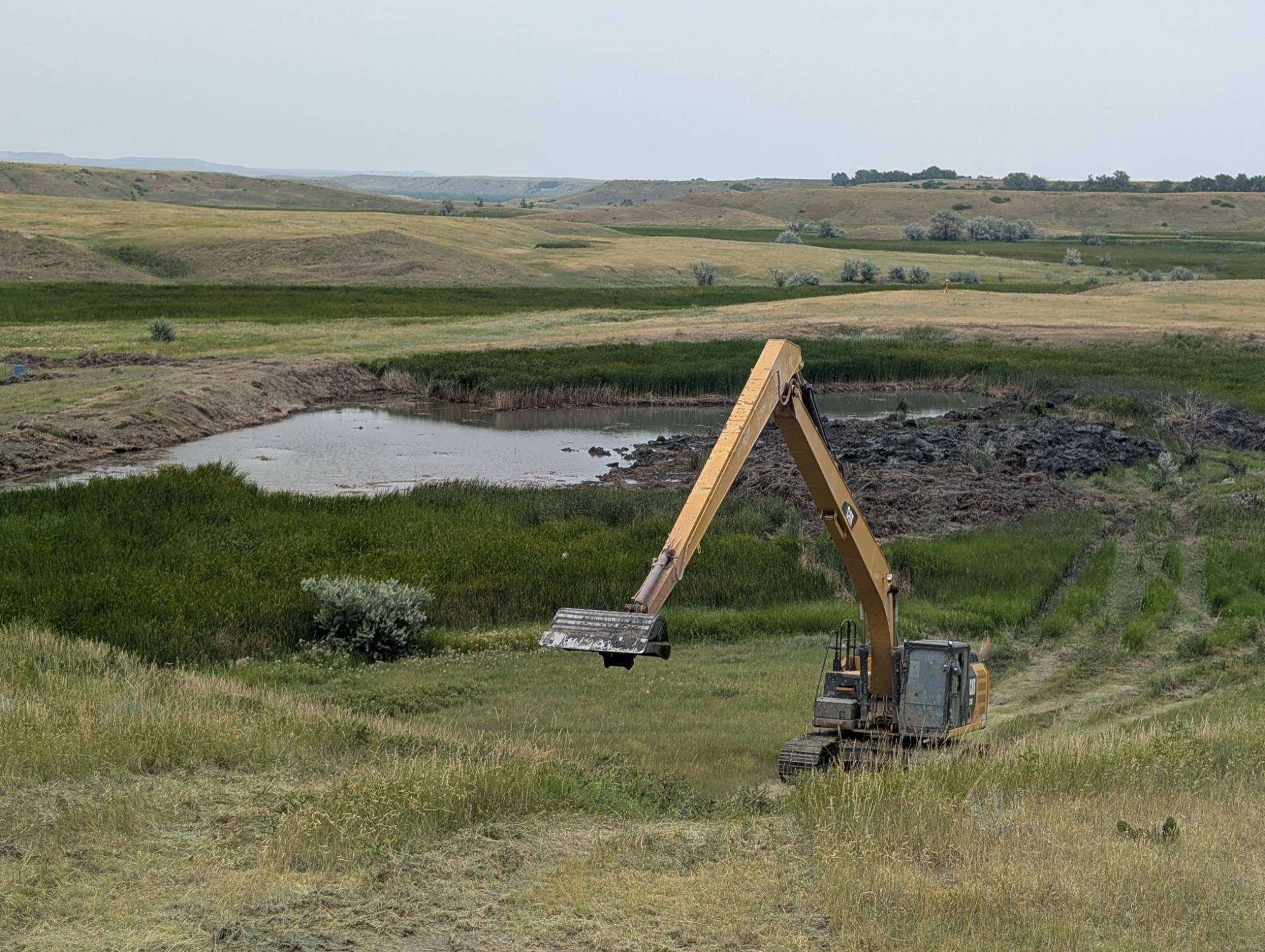 Long-reach excavator above a prairie stock pond partly cleaned of cattails and silt